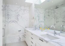 A marble clad drop-in bathtub with a wall mount tub filler is fixed against a marble slab backsplash. A skylight is located over a full-length frameless vanity mirror hung above a white dual washstand finished with flat front cabinets and a marble countertop with polished nickel gooseneck faucets.