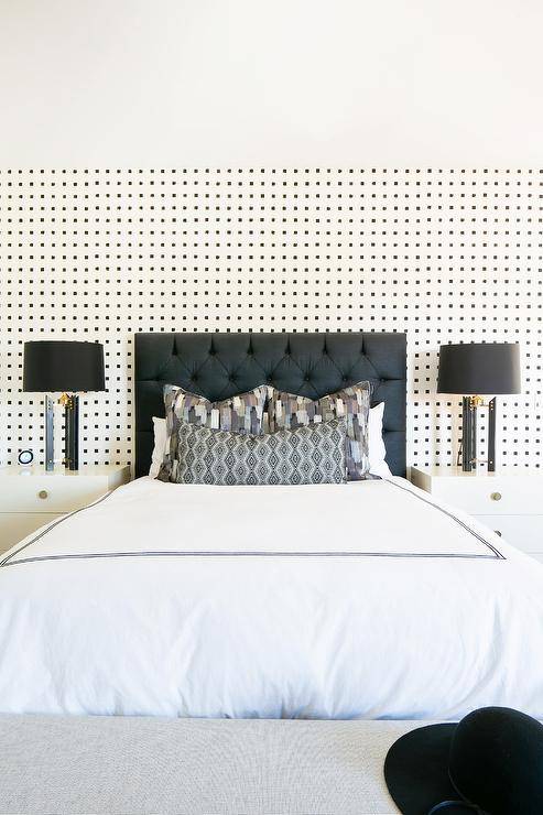 Black and white wallpaper complements a black tufted headboard accenting a bed dressed in black border hotel bedding topped with black and gray pillows. The bed is flanked by white lacquer nightstands illuminated by black lamps.