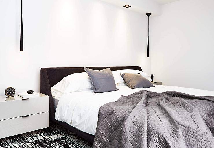 Gray bedding matching gray pillows accents a black wingback bed placed on a black and white rug and matched with white lacquer nightstands lit by black hanging lights.