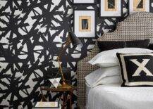 Tan and black in abstract art in black frames hangs from a wall covered in black and white wallpaper over a beige and black houndstooth headboard. The bed is topped with white and black pillows placed on white waffle weave bedding. A black and gold task lamp sits on a gold bedside table beside the bed.