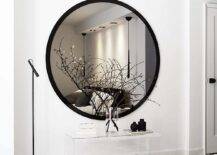 An extra large round black mirror hangs over a lucite bedroom bench placed on a black and white rug lit by a black floor lamp.