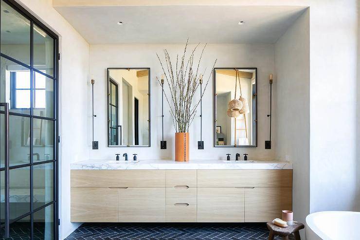 Gorgeous cottage bathroom features black herringbone floor tiles leading to a blond oak double washstand accented with matte black faucets fixed to a honed marble countertop. Black and gold mirrors hang over the washstand and are mounted between long oil rubbed bronze sconces.