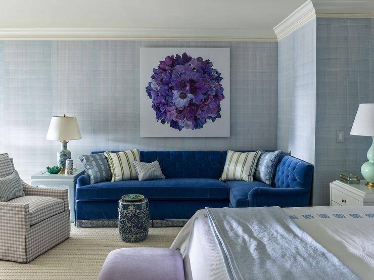 Bedroom features a sitting area with a blue velvet tufted sectional under purple floral art, a blue chinoiserie end table lit by a blue lamp and a gray gingham chair.