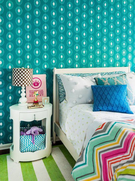 Teal teen girl's bedroom features an ivory bed dressed in green star sheets and a colorful chevron duvet accented with a turquoise trellis print pillow, a blue diamond print pillow, and a teal zebra print body pillow all sat in front of a teal wallpapered wall. Beside the bed, a Bungalow 5 Brigitte 1-Drawer side table holding a teal chevron basket on the bottom and topped with a brown polka dotted four gourd white lamp and a pink canvas print is sat on a striped apple green area rug.