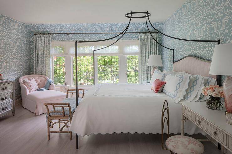 Romantic blue and pink bedroom features walls covered in China Seas' Bali Isle wallpaper accented by matching blue ikat curtains hanging in front of a wall lined with white framed windows allowing natural light to shine on gray wash hardwood floors. An iron canopy bed boasting a pink paisly headboard sits centered on the back wall dressed in white and blue scalloped bedding and completed with a red lumbar pillow. In front of the bed, a blue seat cushion is positioned atop a bamboo bench facing a gray dresser fitted with antique brass pulls and a pink, curved-back, tufted chaise lounge. Flanking the bed, pink marbled glass table lamps decorate a round glass top nightstand a white bamboo desk.