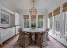 Cottage style dining room with a built-in sideboard accented with a wood top displaying an abstract wall art lit by a set of recessed lights. Ro Sham Beaux Frankie Malibu Chandelier decoratively hangs over a white round dining room table with wicker chairs over a gold and red Persian rug.