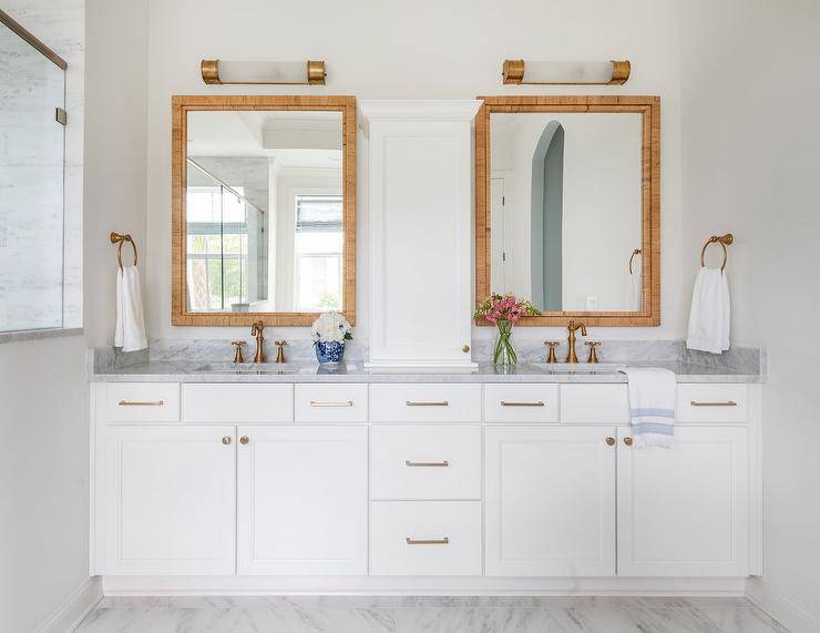 Serena & Lily Balboa Rattan Mirrors hang in a gorgeous bathroom over a white dual bath vanity adorned with brass pulls and aged brass hook and spout faucets fixed to a marble countertop.