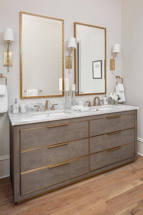 Brass sconces light brass rivet mirrors hung over a gray shagreen double washstand fitted with brass hardware and brass faucets mounted to a marble countertop.