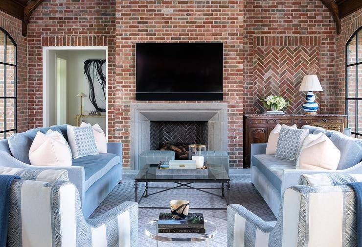 A flat panel television is mounted to a red brick fireplace wall over a gray limestone mantel flanked by a doorway and an antique brown wood cabinet lit by a white and blue lamp and placed against a red brick wall. Light blue velvet sofas accented with white and blue pillows flank a wrought iron coffee table and matched with blue cube stools and blue stripe accent chairs placed on a blue rug.