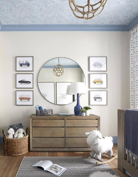 Blue crown molding frames a blue agate wallpapered ceiling in a beautiful nursery featuring vintage car prints hung flanking a round mirror fixed over a brown wooden dresser lit by a blue lamp.