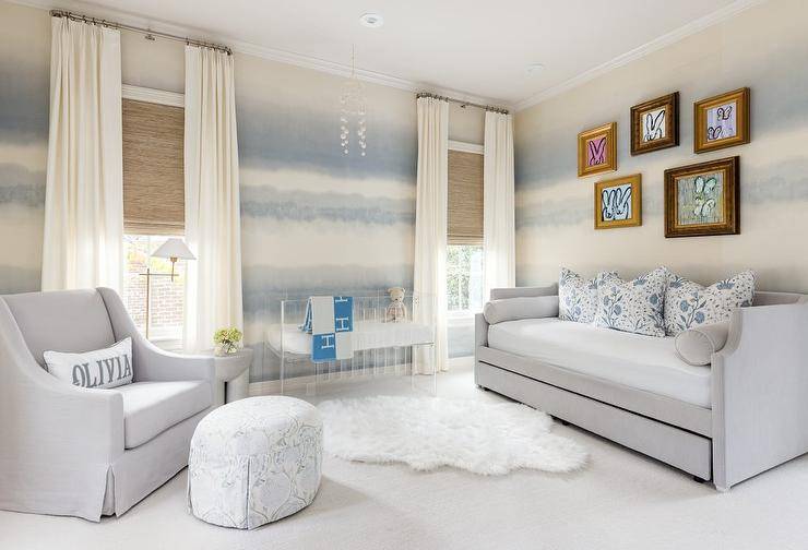 Boy's nursery features a dove gray nursery daybed on white and blue wallpaper with art gallery, a white faux fur rug, a lucite crib with blue Hermes blanket, a dove gray nursery glider with white and blue skirted ottoman and cement accent stool and white curtains.