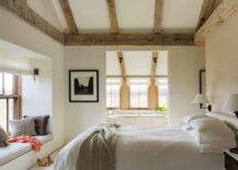 Cottage bedroom boasts a vaulted ceiling accented with rustic wood beams over a taupe headboard on bed dressed in white and taupe bedding flanked by two tone nightstands facing a window seat nook filled with pillows.