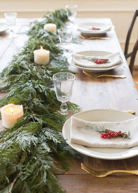 a cozy and homey winter table with an evergreen and pilalr candle runner, berries and evergreens plus gold cutlery