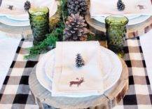 a cozy winter table with a plaid runner, wood slice placemats, large pinecones, green glasses and lots of candles