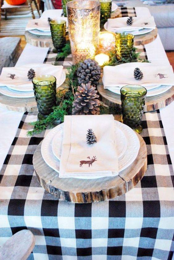 a cozy winter table with a plaid runner, wood slice placemats, large pinecones, green glasses and lots of candles