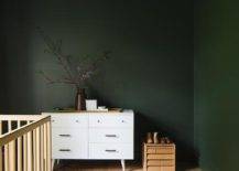 Forest green nursery walls and ceiling complement a white mid-century modern dresser placed behind a pink and blue vintage rug positioned beneath a blond wood crib.