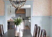 A wood and metal lantern hangs over a brown wood dining table surrounded by vintage wooden dining chairs. Walls half-covered in beige wallpaper are lined with blue crown moldings and finished with blue millwork.