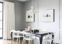 Beautifully designed contemporary dining room is lit by a white faux antlers chandelier hung over a black French dining table surrounded by Kartell Masters Chairs placed on a wood herringbone floor. Blue art hangs on a gray wall accented with gray paneling and gray crown molding, while white French pleated curtains hang in front of black glass doors.