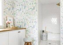 Clad in floral wallpaper, this welcoming bathroom features a doorless walk-in shower covered in white subway surround tiles and fitted with a subway tiled bench fixed behind a white and blue pierced drum stool. A wooden stool sits outside of the shower and in front of white cabinets topped with a brown wood countertop mounted beneath a window.