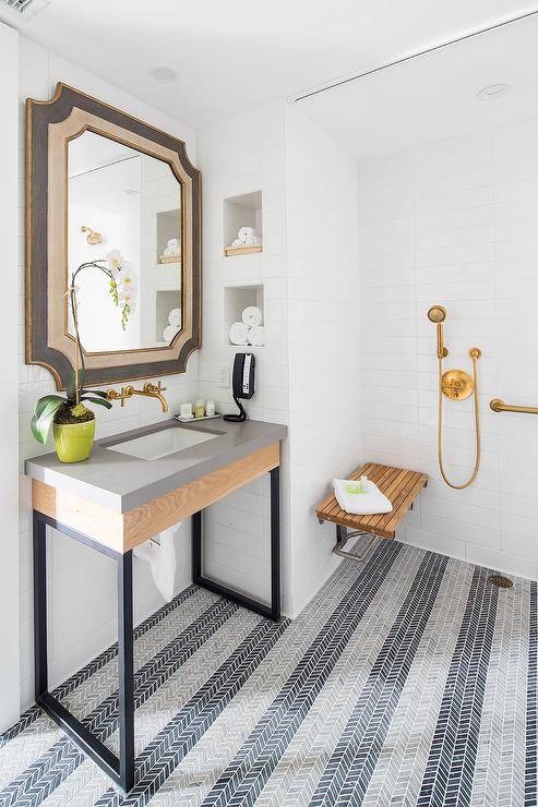 Rectangular bathroom floor tiles laid in a herringbone pattern is a look that many can't get enough of. Here, gray and blue floor tiles create a stunning shower design in an open shower concept with a teak bench and a brushed gold kit. This doorless shower displays stacked white shelves beside the vanity space showcasing a gray and gold French mirror over a wood and metal sink vanity.