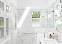 Exquisite bathroom with marble tiled floors leading to a marble tiled walk-in shower. The shower features a small window with frosted glass panes pairing with a skylight overhead which fills the space with natural light. A marble tiled shelf within the shower adds storage and display space. The modern chrome based washstand with white counter, pairs with a pivot wall mirror flanked by polished nickel and frosted glass sconces. Built-in cabinetry with glass fronted upper cabinets add plentiful storage to the space. Vaulted beadboard ceilings complete the space.