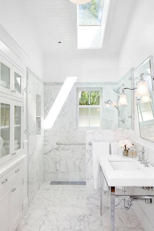 Exquisite bathroom with marble tiled floors leading to a marble tiled walk-in shower. The shower features a small window with frosted glass panes pairing with a skylight overhead which fills the space with natural light. A marble tiled shelf within the shower adds storage and display space. The modern chrome based washstand with white counter, pairs with a pivot wall mirror flanked by polished nickel and frosted glass sconces. Built-in cabinetry with glass fronted upper cabinets add plentiful storage to the space. Vaulted beadboard ceilings complete the space.