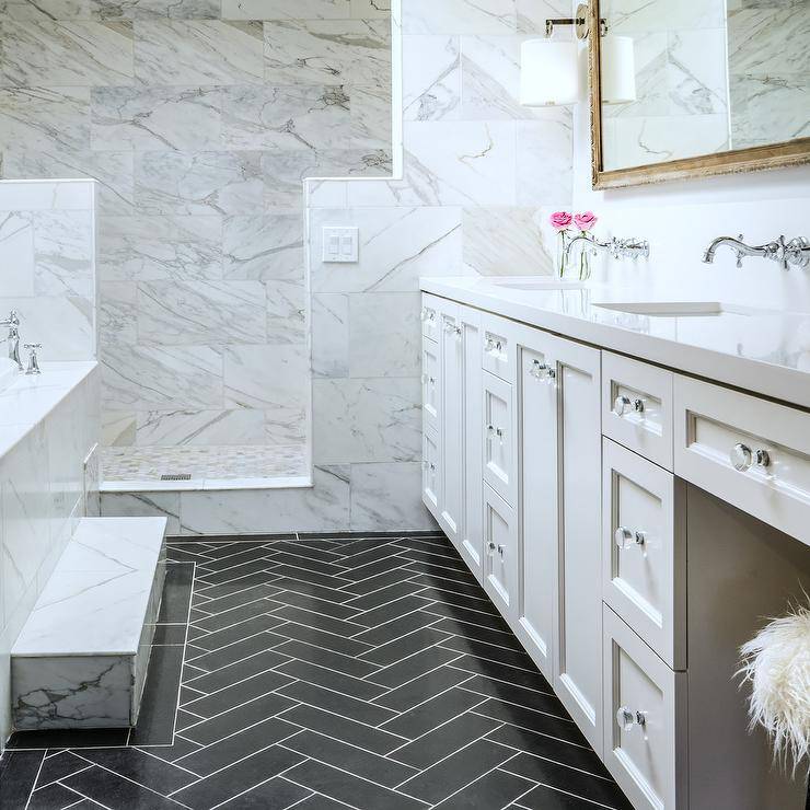 Barbara Barry French Cuff Sconces light up a marble bathroom flanking a French wood vanity mirror over a white dual washstand with white quartz counters. Clean, white sinks fitted with wall mount hook and spout faucets pair with white shaker cabinets and lucite glass knobs. The result is a master bathroom that feels elegant yet inviting with a bit of dark tones from the black herringbone floor tiles. A marble step to bathtub walk-in shower is doorless allowing the inner marble tiles to display without distraction.