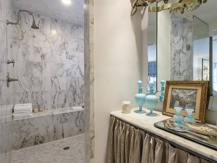 Beautiful shower boasts gray and white marble tiled surround and marble clad built-in shower bench with gooseneck shower head over diamond marble mosaic tiled shower floors. The entryway leading to the walk-in shower features a burlap skirted dressing table with marble counter topped with a mirrored tray, seaglass glassware and a leaning oil-painting resting against a frameless vanity mirror.