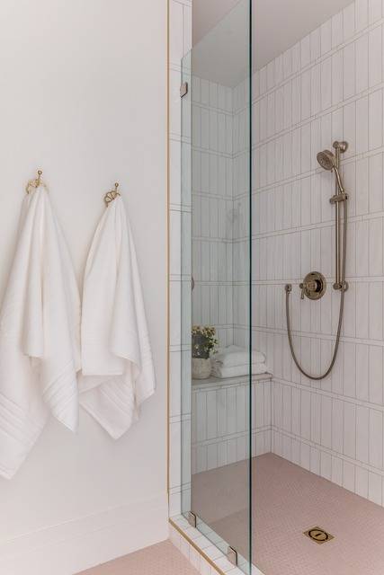 Shower features pink floor tiles, white vertical stacked shower tiles with a satin nickel shower kit, a built in shower bench and a glass partition.