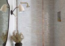 A glass partition with brass trim accents a shower clad in gray glazed surround tiles framing a niche located over a black stool. The shower is finished with a brass shower kit.