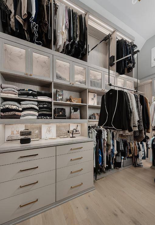 Gray walk in closet features custom drop down clothes racks, a built in dresser with brass pulls under gray shelves and a tall ceiling.