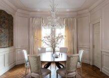 Elegant French dining space boasts a crystal chandelier fixed over a round dining table surrounded by pink and green damask French dining chairs positioned on a herringbone wood floor. Pale beige wainscot walls are lined with matching crown molding, while windows are covered in light beige curtains.