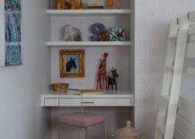 A lucite chair on wheels sits in a girl's bedroom at a white floating desk mounted in a nook against walls clad in purple lotus print wallpaper. Styled white floating shelves are fixed above the desk and over a gold framed art piece.