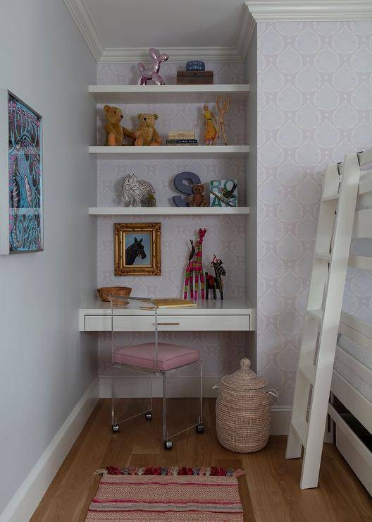 A lucite char on wheels sits in a girl's bedroom at a white floating desk mounted in a nook against walls clad in purple lotus print wallpaper. Styled white floating shelves are fixed above the desk and over a gold framed art piece.