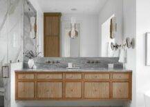 Marble floor tiles are fixed beneath a brown floating dual washstand adorned with brass knobs and a gray marble countertop. A brass faucet kits are fixed to a gray marble backsplash beneath a frameless vanity mirror finished with three brass and alabaster sconces.