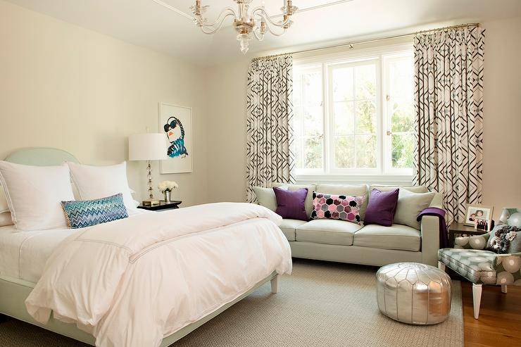 Contemporary teenage girl's bedroom features a silver metallic Moroccan pouf sat catty corner on a gray diamond print rug in front of a black and gray printed accent chair located beside a gray sofa topped with aubergine pillows and accented with a square black side table. The sofa is positioned under a window covered in gray and pink diamond print curtains lit by a glass chandelier. A mint green camel-back bed dressed in white hotel bedding complimented with a blue chevron lumbar pillow is placed beside a black nightstand lit with a stacked glass ball table lamp next to a framed print.