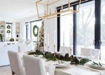 Dining room features a Christmas garland and acorn runner on a long off white dining table with off white fabric dining chairs illuminated by a Darlana rectangular linear lantern.