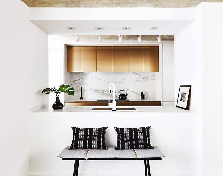 A black bench with gray cushions and black and white striped pillows sits against the back of a kitchen pass through boasting a sink white a white modern gooseneck faucet. Gold cabinets are mounted against a marble slab backsplash over an induction cooktop.