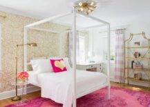 Pink and gold teenage girl's bedroom features a brass sputnik flush mount, Jonathan Adler Sputnik 12 Light, placed over a white canopy bed dressed in white bedding and a pink and gold metallic heart pillow lining an accent wall clad in gold metallic wallpaper, Oh Joy Petal Pusher Wallpaper, atop a hot pink overdyed rug. A white canopy bed is flanked by an acrylic tulip table and a brass floor pharmacy lamp to the left and a white desk with Ghost Chair to the right. Chic girl's bedroom boasts a gold metal bamboo etagere placed next to windows dressed in pink striped curtains layered over white roman shades.