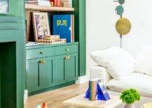 Green shaker lacquered living room cabinets adorned with brass knobs and fitted beneath inset blond wood floating shelves mounted against a mirrored backsplash and beside a green art deco fireplace. Staggered blond wood tables sit on a rug in front of a white sectional.