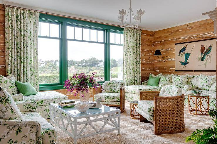 Welcoming beige and green cottage living room features green framed windows covered in green chintz curtains and framed by a wall clad in faux bois wallpaper. Green chintz sectionals are placed facing each other on a woven rug, while a white rope coffee table sits between one sectional and two barrel back cane chairs with green chintz cushions.