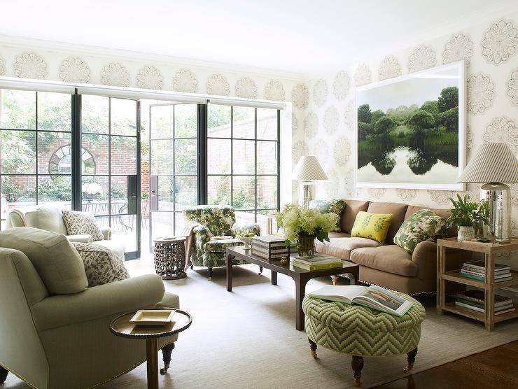 A brown and green living room brings a traditional design while feeling fresh and updated. Here, a living room is furnished with a brown roll arm sofa with green pillows facing a black coffee table and round green chevron tufted ottoman. An outdoor lake wall art decorates beige print wallpaper walls above the sofa complementing a palm leaf print accent chair and green decor. Black frame glass doors feature a stunning view into an outdoor patio uniting the outdoors with indoor charm.