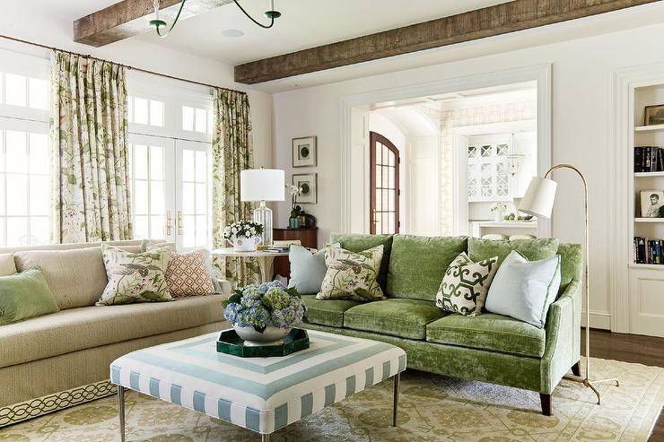 Beneath a ceiling accented with rustic wood beams, a green velvet sofa topped with blue and green pillows placed on a beige and green rug facing a blue concentric ottoman. Botanical print curtains cover French doors and windows located behind a tan sofa.