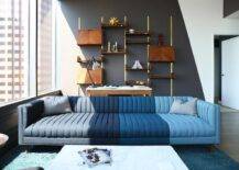 High rise apartment boasts a wonderfully designed contemporary living room featuring a blue channel tufted sofa placed on a blue rug in front of a desk positioned against brass and wood ladder shelves finishing a white and black painted accent wall.