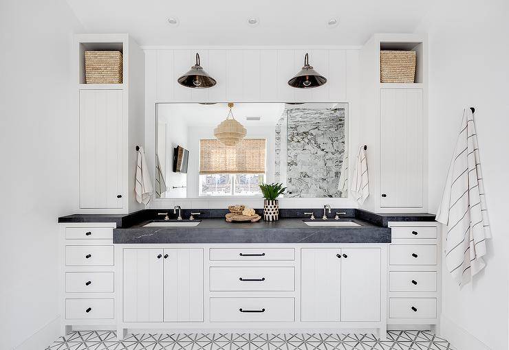 White and gray lattice floor tiles lead to a white plank double washstand adorned with oil rubbed bronze hardware and a thick honed black marble countertop. Sink with polished nickel faucet are located under a frameless vanity mirror lit by two vintage polished nickel lanterns mounted to a vertical plank backsplash. The lanterns are flanked by cubbies holding woven bins placed over linen cabinets.