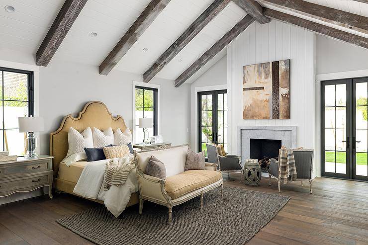 Gold French bed with a French settee at the foot atop a black jute rug in a transitional bedroom design. Crystal lamps on gray French nightstands add a luxurious look flanking a well-adorned bed. Rustic wood ceiling beams add a cottage appeal to the white vaulted ceiling over the bedroom completed with a vertical shiplap fireplace wall.