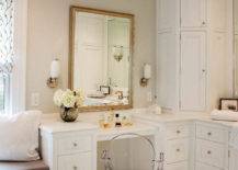 Stunning bathroom with built-in window seat to the left of a next to a drop-down vanity lined with a Ghost Chair. A large gilt mirror flanked by nickel and white glass sconces hangs above the vanity with a corner cabinet finishing the space over marble tiled floors.