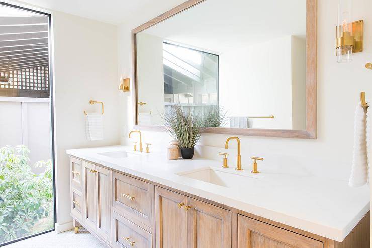 A brown brushed oak dual washstand adorned with brass hardware is topped with a white quartz countertop holding brushed gold faucets beneath a brown brushed oak vanity mirror flanked by brass and glass sconces.