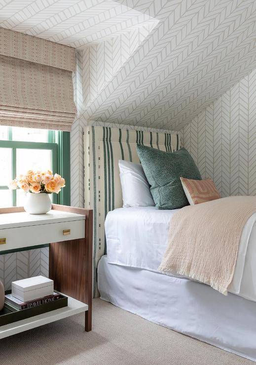 Attic bedroom features a white and brown nightstand accented with brass hardware and an an ivory and green striped headboard accented with tan and green pillows on Serena & Lily Feather Wallpaper.