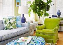 Contemporary green and blue living room features an oval green and blue tufted ottoman placed in front of a blue striped sofa accented with blue and green pillows. Stacked art pieces hang from a green wall over a white Parsons side table illuminated by a cobalt blue lamp, as a fiddle leaf fig plant sits next to a green velvet roll arm chair.